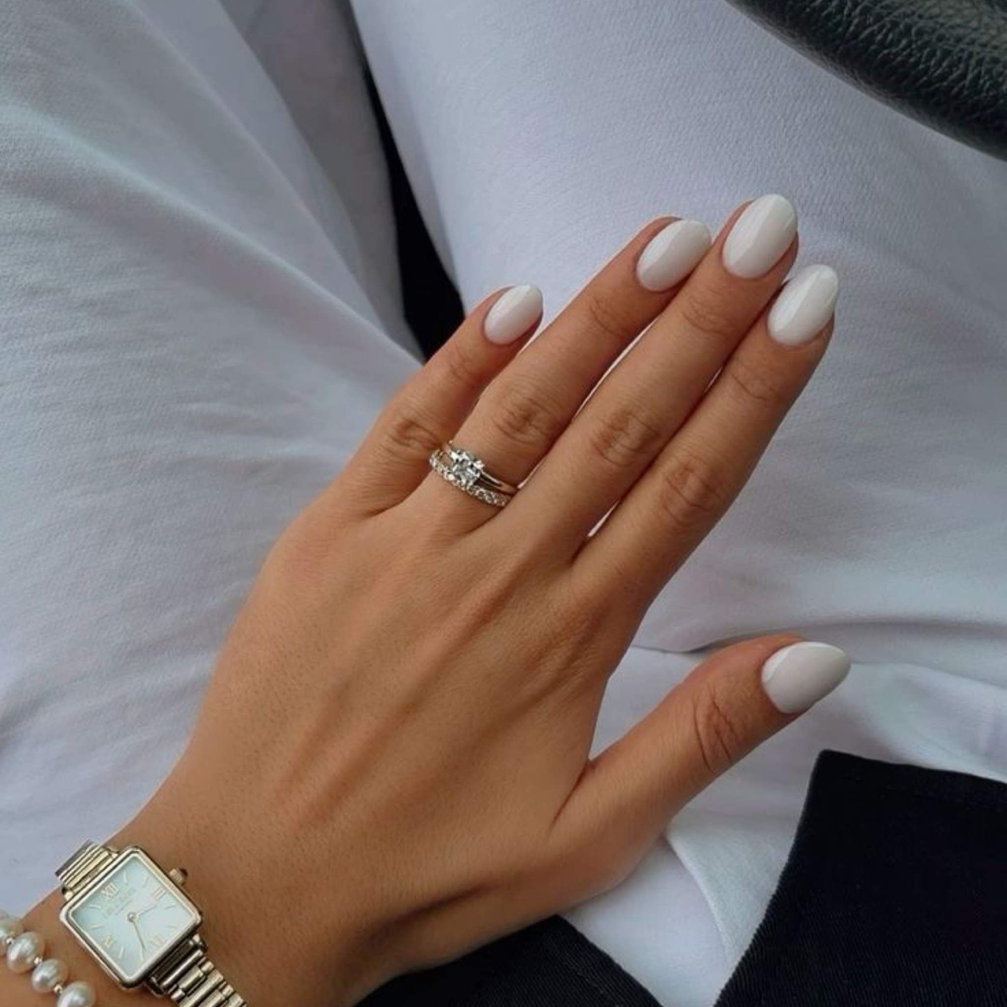 Hand with white nail polish wearing a ring and watch, against a neutral background